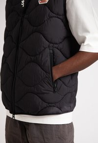 Svart vadderad väst med ett vågmönster, med zippered fickor och hög krage, lager-på-lager över en vit skjorta. Strukturerad tygdetalj synlig.