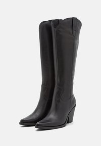 Botas altas de cuero negro hasta la rodilla con punta afilada y borde ondulado. Presentan un tacón apilado y detalles de costura mínimos.