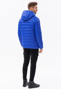 Giacca a vento blu con cappuccio, caratterizzata da un design trapuntato, texture liscia e polsini a coste, abbinata a pantaloni cargo neri e stivali.