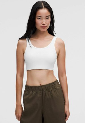 Giovane donna con lunghi capelli neri che indossa un top corto bianco e pantaloni larghi verde scuro, in piedi davanti a uno sfondo chiaro e uniforme.