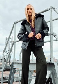 Giacca grigia con zip e colletto e polsini in pelliccia sintetica, abbinata a pantaloni a gamba larga verde scuro. Il modello si trova su una struttura metallica.