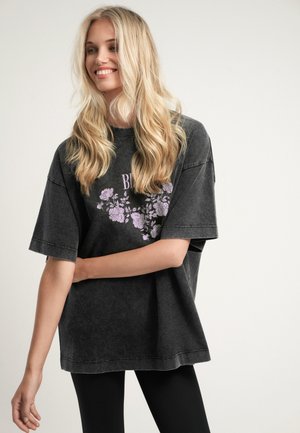 Blonde vrouw glimlachend, draagt een oversized vervaagd zwart T-shirt met paars bloemmotief en zwarte legging, staand tegen een eenvoudige achtergrond.