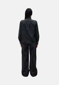 Person mit langen schwarzen Haaren trägt eine passende schwarze Denimjacke und weite Jeans, steht mit dem Rücken zu einem schlichten Hintergrund.