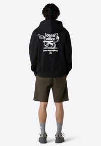 Černá mikina s kapucí a bílým grafickým potiskem na zadní straně, obsahující text a ilustraci. V kombinaci s olivovými kraťasy a šedými ponožkami.