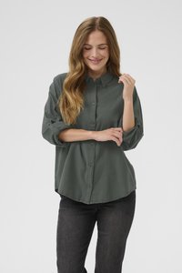 Žena s dugom valovitom kosom, odjevena u tamnozelenu košulju na kopčanje i crne traperice, smiješi se s zatvorenim očima na jednobojnoj bijeloj pozadini.