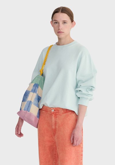 Jeune personne portant un sweat-shirt bleu clair et un pantalon orange, avec un sac à carreaux bleu et crème, à bandoulière jaune, porté sur une épaule.
