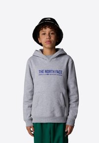Felpa con cappuccio grigia e tasca frontale, con testo blu "THE NORTH FACE" e logo. Indossata con pantaloni verdi e un cappello a secchiello nero.