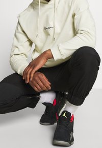 Felpa colore beige chiaro con cappuccio e logo nero, abbinata a joggers neri con cerniere e sneakers nere con dettagli rossi.