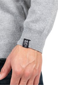 Cuff di maglione grigio a maglia con texture a coste. Presenta un'etichetta in tessuto nera con un personaggio di teschio a cartone animato e un dettaglio a cuore rosso.