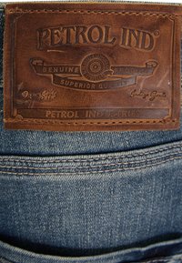 Ruskea nahkalaatta, jossa on kohokuvioitu "PETROL IND" -logo, kiinnitetty sinisiin farkkuihin, joissa on näkyvät tikkaus ja tekstuurinen pinta.