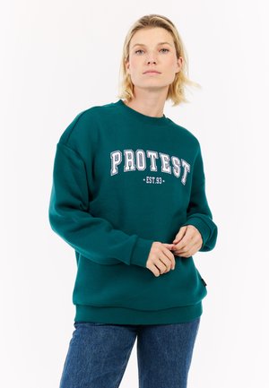 Jonge vrouw met kort blond haar in een teal sweatshirt met "PROTEST EST. 93" in witte letters en blauwe jeans tegen een witte achtergrond.