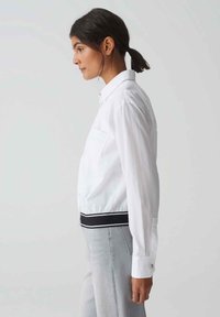 White cropped shirt with long sleeves, featuring a black and white striped waistband and a smooth texture. Side view shows one chest pocket.