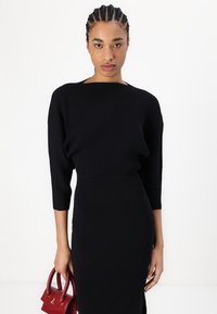 Pull noir à côtes avec un décolleté bateau et des manches trois-quarts, associé à une robe noire ajustée et un petit sac à main rouge.