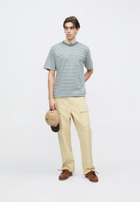 Ριγέ μπλουζάκι σε μπ λε και λευκό, μπεζ παντελόνι cargo, καφέ παπούτσια, κρατώντας ένα καφέ καπέλο. Καθημερινό σχέδιο με χαλαρή εφαρμογή και λεπτομέρειες στις τσέπες.