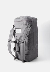 Sac à dos gris de forme rectangulaire, avec des bretelles rembourrées, une poche zippée à l'avant et une fenêtre transparente pour carte d'identité. Matériau durable et coutures renforcées.