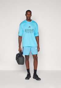 Gaiši zila sporta krekls ar melno "ADIDAS" logotipu, kombinēts ar saskaņojošām šortiem. Modelis tur melnu sporta somu un valkā melnus sporta apavus.
