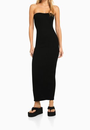 Femme portant une robe maxi noire sans bretelles, ajustée, associée à des sandales plateformes noires, sur fond blanc.