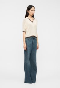 Haut en maille à texture crème avec un col en V et bordure marron, associé à un jean denim bleu à jambes larges et lavage moyen.