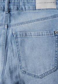 Jeans de denim azul claro con un bolsillo trasero de forma rectangular, un desvanecimiento sutil y costuras contrastantes. Etiqueta visible en la cinturilla.