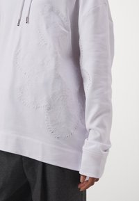 Sweat à capuche blanc avec des motifs complexes de broderie à œillets sur le devant et la manche, porté avec un pantalon gris foncé sur un fond uni.
