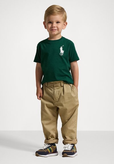 Grön t-shirt med vit logotyp, beige baggy byxor och svarta sneakers med gula och gröna accenter. Barnet står med ett neutralt uttryck.