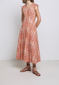 Punainen ja kerma värinen kuviollinen mekko, jossa on V-kaula-aukko ja solmittavat olkaimet. Mekossa on virtaava A-linjainen siluetti ja polvipituinen hame. Ruskeat narukengät.
