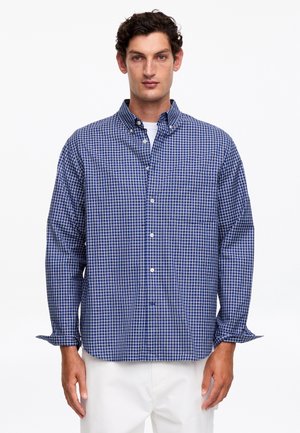 Mann trägt ein blau-weiß kariertes Langarmhemd über einem weißen Unterhemd und weiße Hosen vor einem einfarbigen Hintergrund.