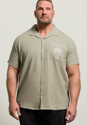 Muskeløs mand iført en lyseolivengrøn kortærmet skjorte med knapper og palmebladbroderi på brystlommen mod en ensfarvet baggrund.