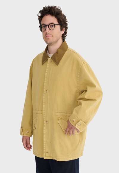 Uomo con capelli ricci e occhiali che indossa una giacca leggera marrone chiaro, ampia, con polsini con bottoni e tasche frontali inclinate, su uno sfondo semplice.