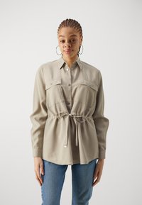 Light beige shirt with two chest pockets, button closure, adjustable waist tie, and long sleeves, paired with blue jeans.