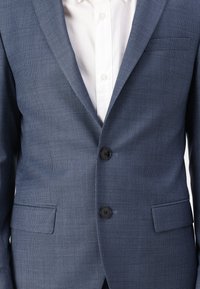 Blauer Blazer mit Muster aus strukturiertem Stoff, ausgestattet mit zwei schwarzen Knöpfen, Reverskragen und einer einzelnen Brusttasche. Darunter ein weißes Hemd.
