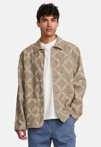 Beige jacket with abstract pattern, button closure, two front pockets, and a relaxed fit, worn over a white shirt.