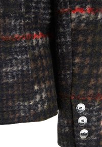 Villa sekoitettu takki, jossa on tumman ruudullinen kuosi vihreillä, ruskeilla ja punaisilla yksityiskohtilla. Kolme metallista nappia ranteessa tuo esiin tekstuuria.