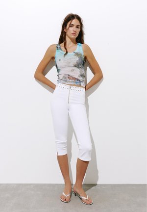Femme portant un crop top imprimé sans manches, un pantalon court blanc taille haute avec des clous, et des sandales blanches à talons, debout contre un mur blanc.