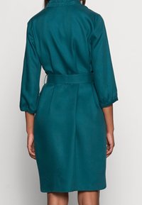 Turkis kjole med bælte i taljen, trekvartlange ærmer og glat tekstur. Simpelt design med krave og knælængde.