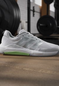 Chaussure de sport blanche avec détail vert sur la semelle, placée sur le sol de la salle de sport avec du matériel de musculation en arrière-plan flou.