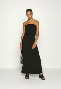 Frau mit geflochtenem Haar, die ein trägerloses schwarzes Maxikleid, schwarze High Heels, goldene Ohrringe, eine Halskette trägt und eine schwarze Handtasche hält.
