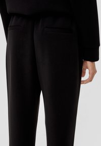 Zwarte stoffen broek met een gladde textuur, voorzien van twee discrete pockets aan de achterkant en een ontspannen pasvorm.