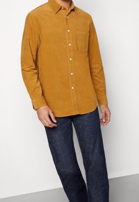 Senapsgul korduléjskjorta med knappkrage, en bröstficka och vita knappar, kombinerad med mörkblå jeans.