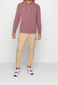 Man wearing mauve hoodie, beige cargo pants with side pockets, and white sneakers with purple and neon green accents, standing against a plain background.