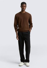 Brauner Strickpullover mit Rundhalsausschnitt, kombiniert mit schwarzer Hose und weißen Sneakers. Glatte Textur, entspannte Passform, minimalistische Details.