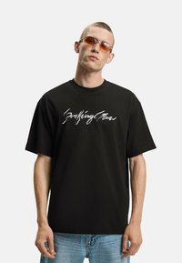 T-shirt in cotone nero con scritte bianche sul davanti, abbinato a occhiali da sole con tonalità arancione. Maniche corte e vestibilità ampia.