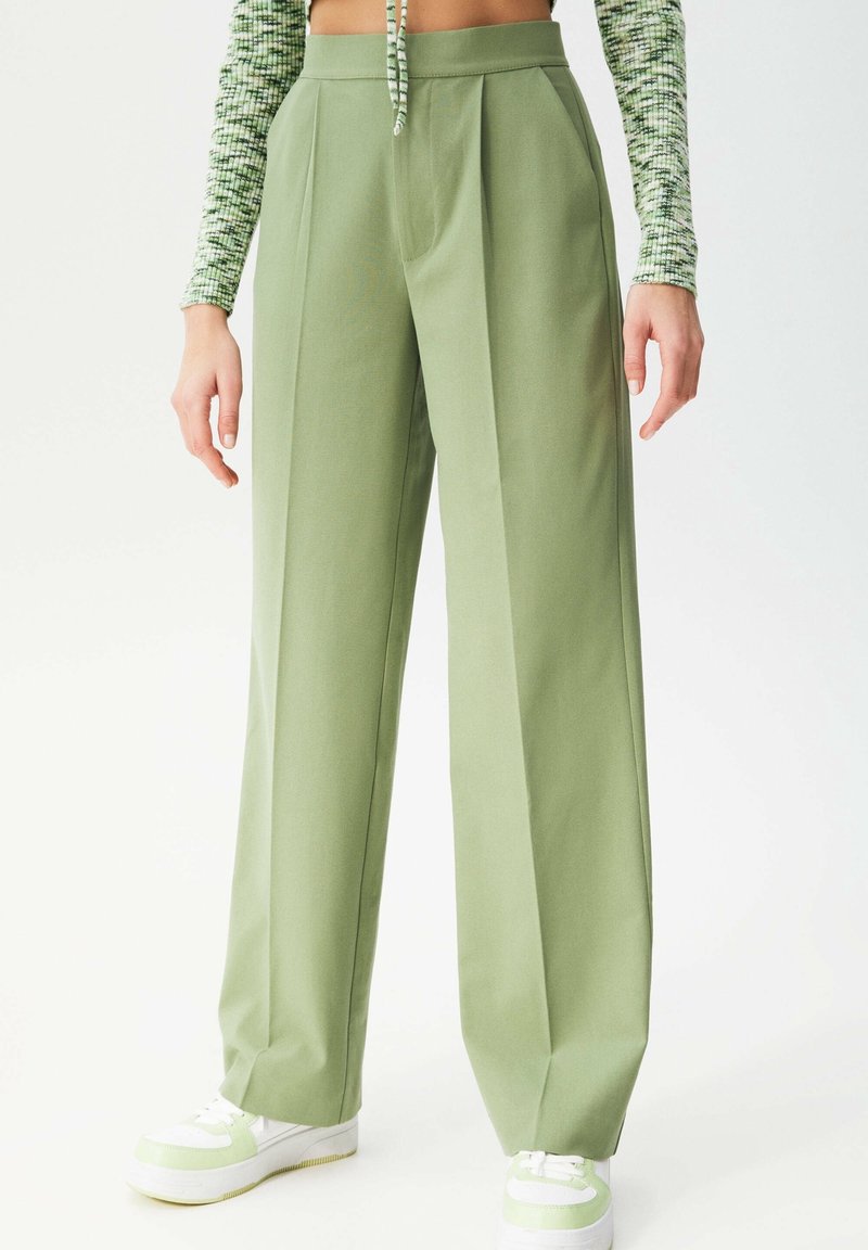 Pantalon vert clair à taille haute, jambes larges, et plis devant. Porté avec des baskets blanches ornées d'accents verts. Texture de tissu lisse.
