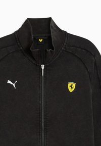 Sort zip-up jakke lavet af blødt stof med ribbet krave. Har et hvidt Puma-logo og et gult Ferrari-våbenskjold med røde og grønne accenter.