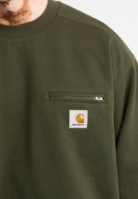 Sudadera de algodón verde con cuello redondo, un bolsillo en el pecho con cremallera y un parche cuadrado de logo de Carhartt en amarillo y blanco. Textura suave.