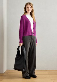 Purple ribbed cardigan with black buttons, worn over a white shirt, paired with high-waisted gray trousers and a black leather tote bag.