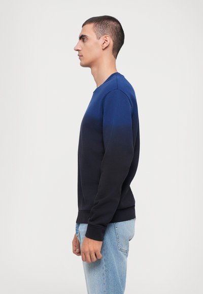 Sweatshirt bleumarin cu o degradare a culorii către negru, decolteu rotund și manșete ribbate. Modelul poartă blugi de culoare albastru deschis.