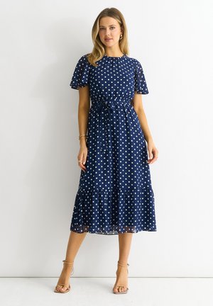 Navy blue polka dot dress with short flutter sleeves, a cinched waist, and a tiered ruffled hem. Made from lightweight, flowy fabric.