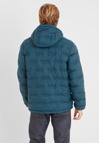 Teal puffer jacket with a hood, featuring a quilted design and elastic cuffs. Worn over dark denim jeans, showcasing soft, insulated material.