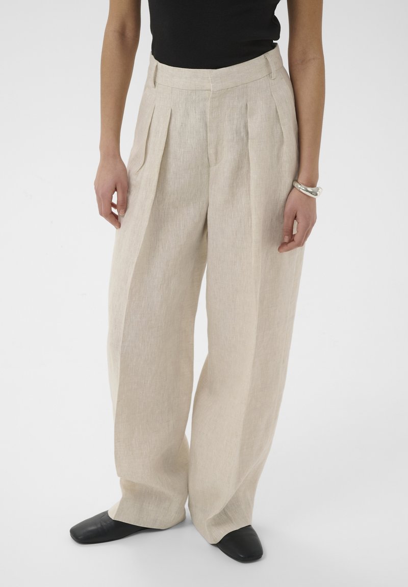 Femme portant un pantalon beige évasé plissé, un haut noir sans manches, des chaussures noires et un bracelet argenté au poignet droit.